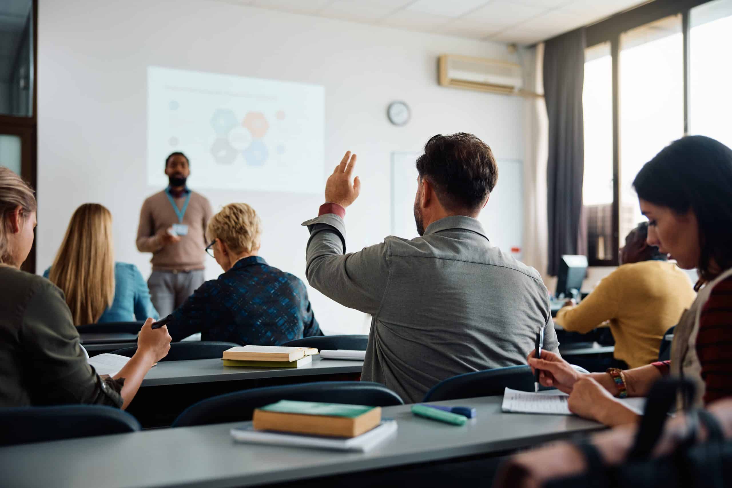 Back view of older student raising his hand to answer teacher's question during education training class.