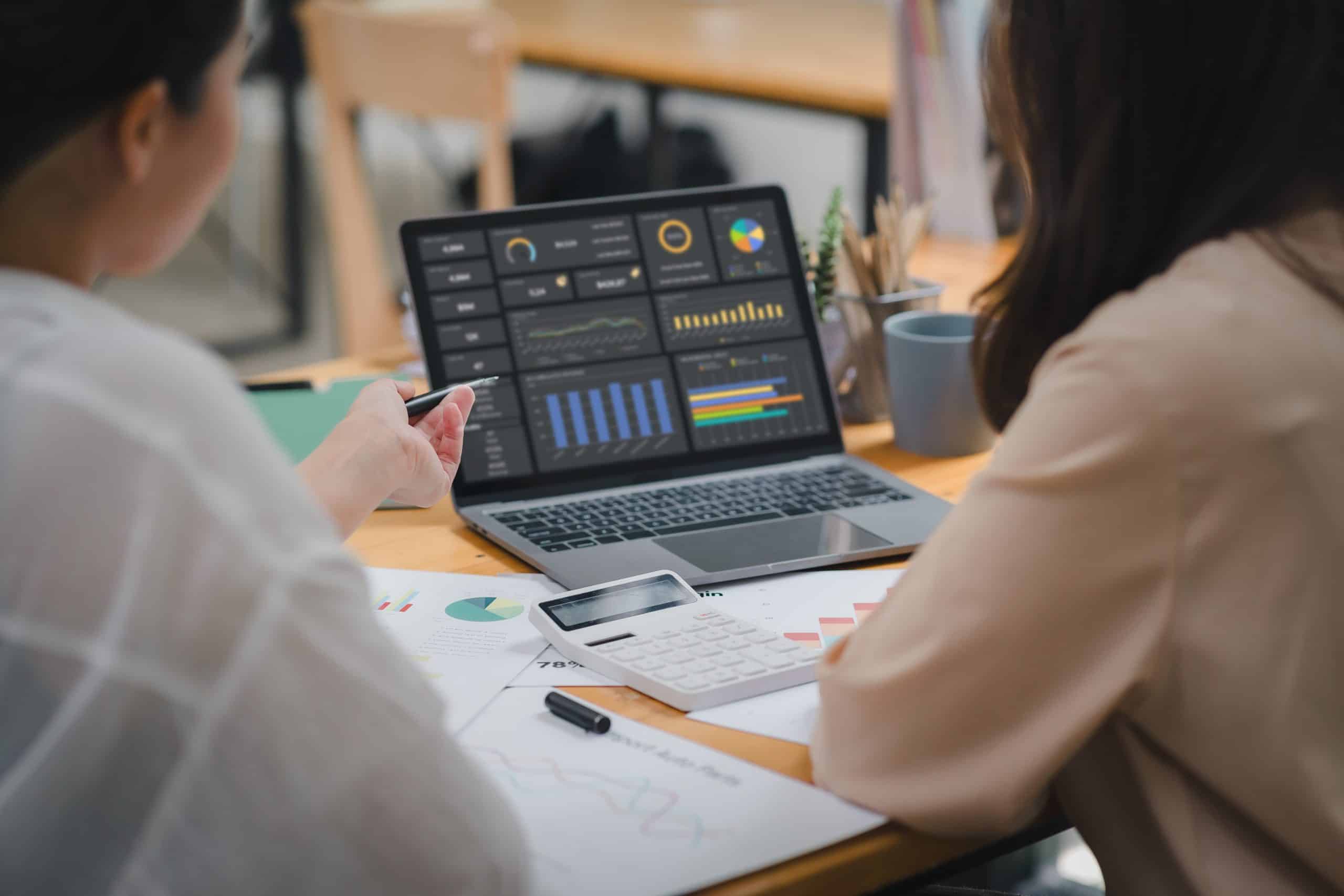 Two businesspeople or an accountant team are analyzing data charts, graphs, and a dashboard on a laptop screen in order to prepare a statistical report and discuss financial data in an office.